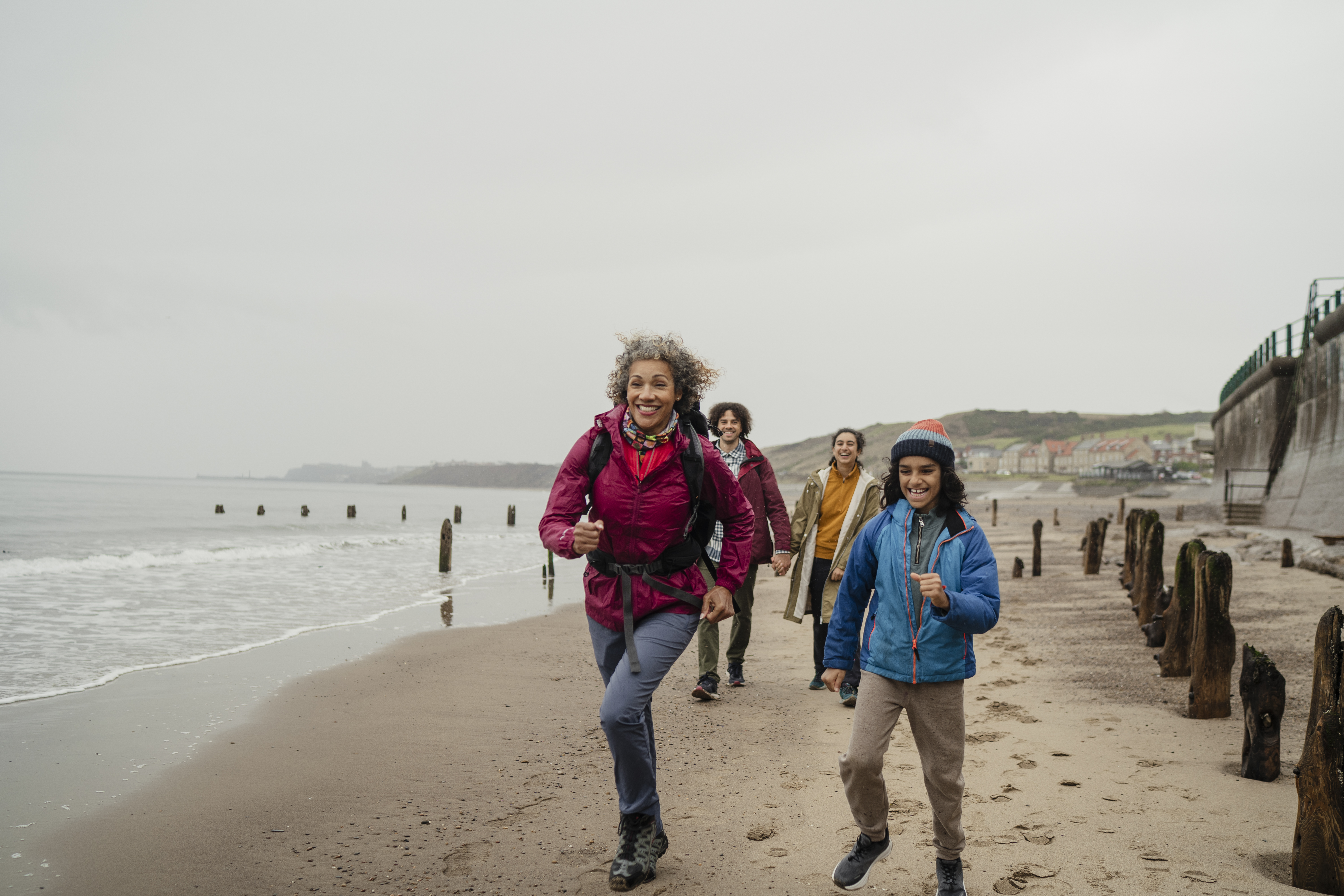 Group of people hiking and smiling along a sandy beach with wooden groynes, some houses, cliffs, and sea in the background on a cloudy day.