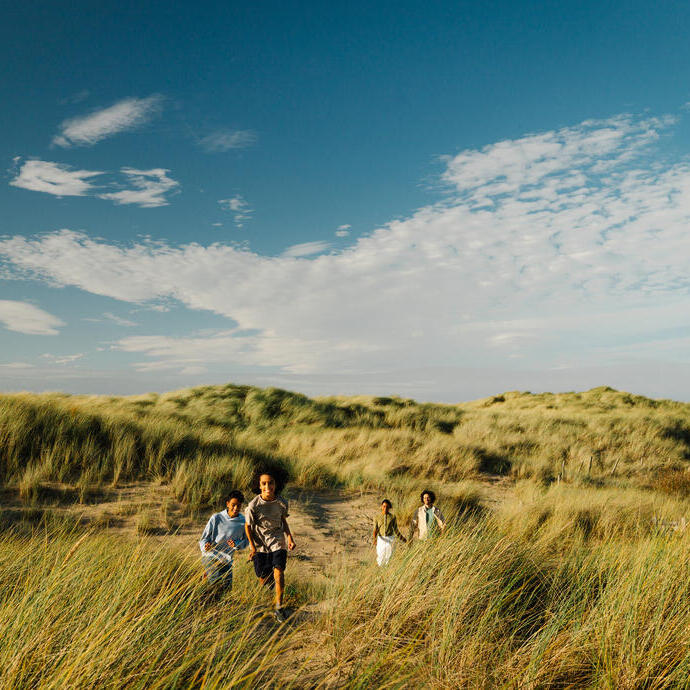 Children running through grassy sand dunes under a bright blue sky with scattered clouds.