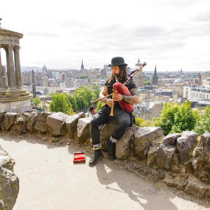 Dudelsackspieler sitzt auf einer Steinmauer mit Stadtblick und klassischem historischen Denkmal links.