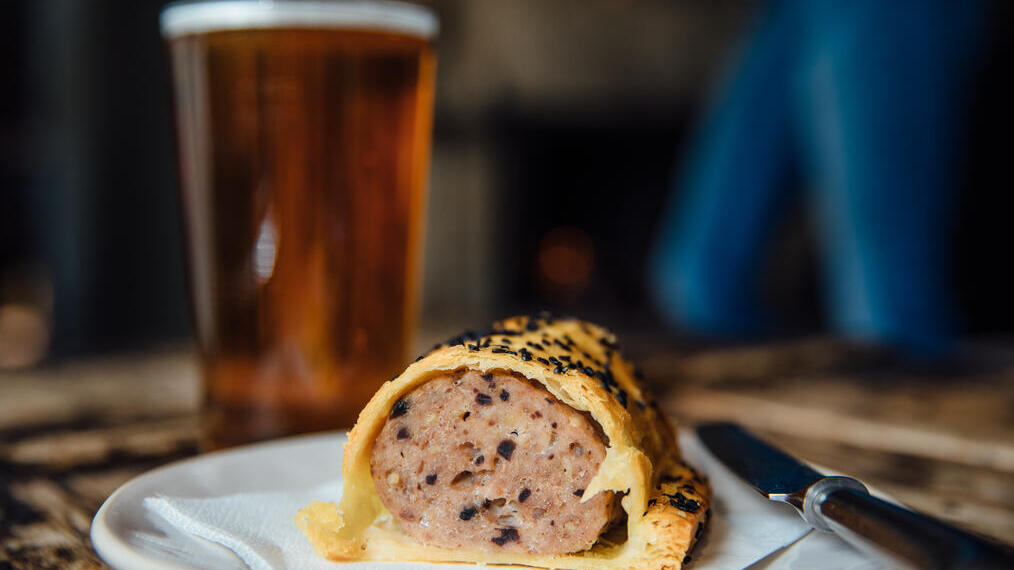 Pint of beer and a sausage roll on a plate on a table in a pub