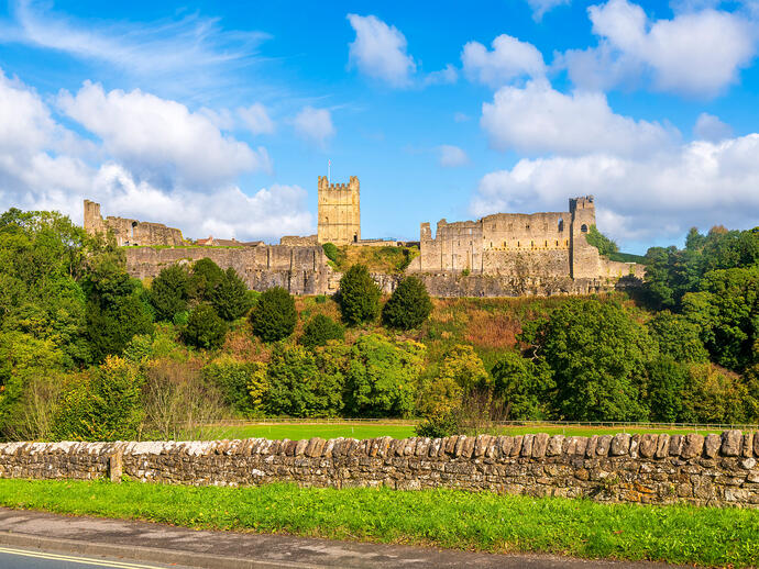 Historic stone castle ruins on a hill with green trees and a stone wall under a blue sky with scattered clouds.