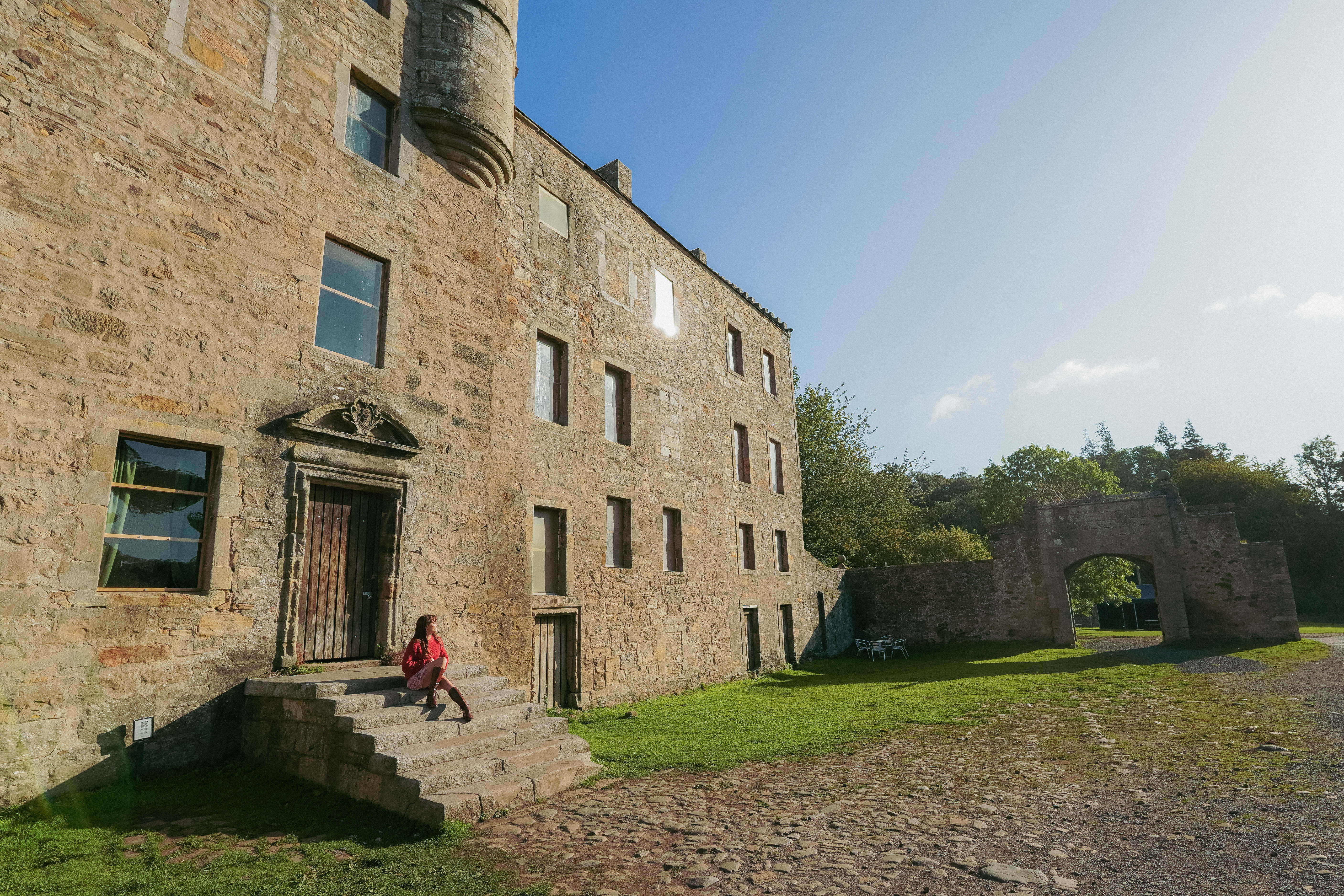 Historic stone building with arched entry gate, green lawn, and cobbled path. A person sits on steps in afternoon sunlight.