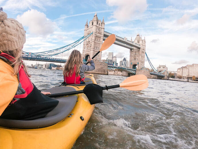 Two women paddling in a kayak on a river towards a bridge in a city