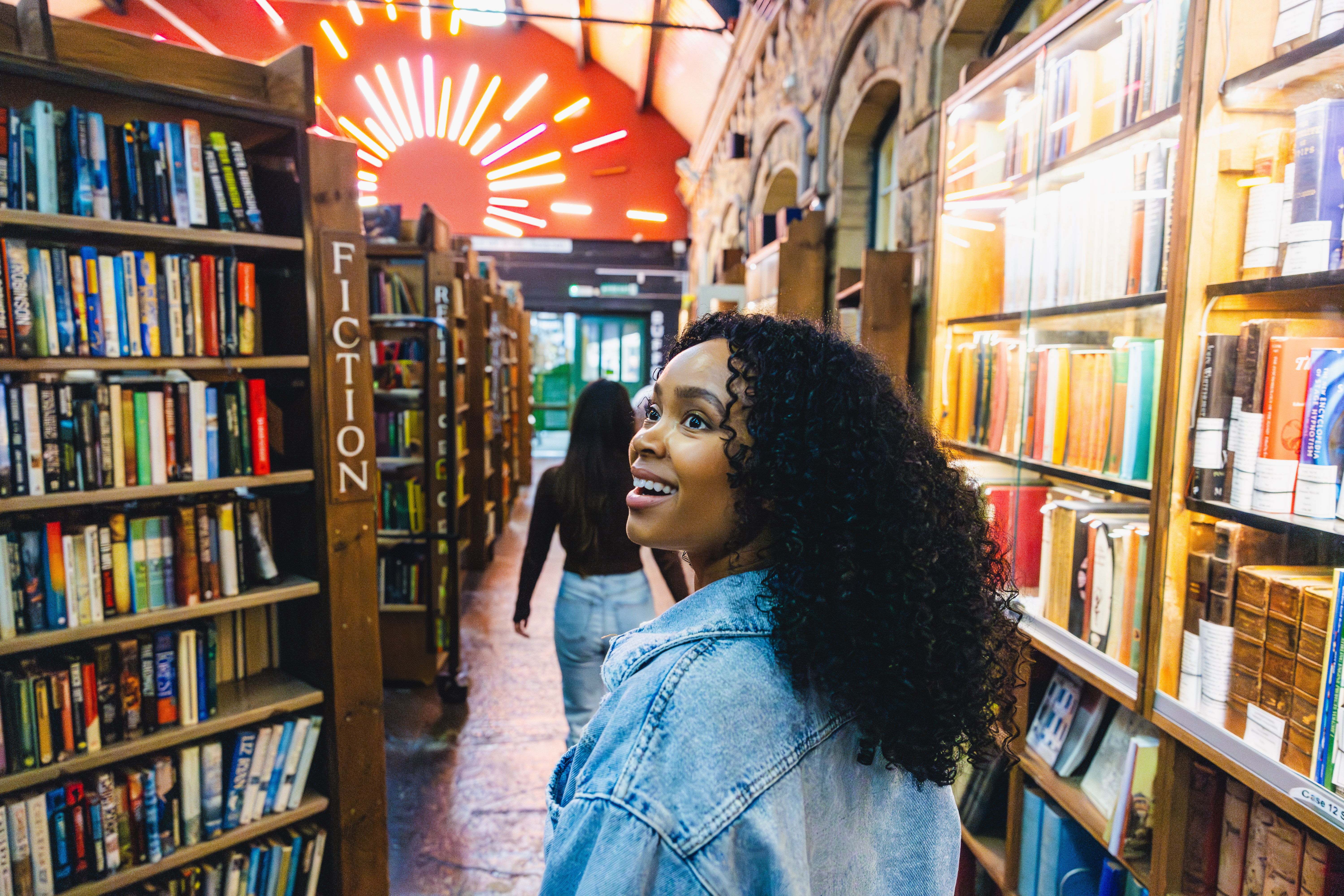Two people walk through a vibrant, well-lit bookstore with shelves of books, stone walls, and neon lights overhead.