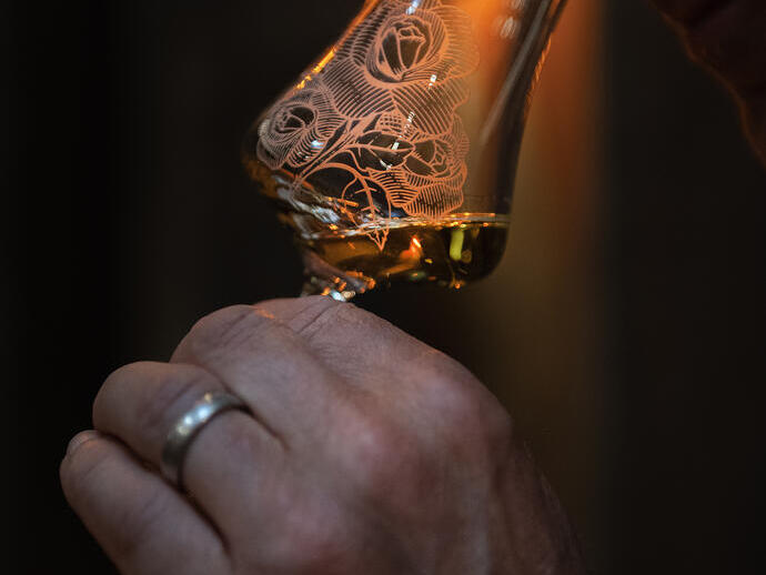 A person smelling a patterned whisky glass with amber liquid, highlighted by warm light in a dark setting, evoking tasting or sensory evaluation.