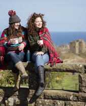Two women with hot drinks, sitting on a stone wall near a castle