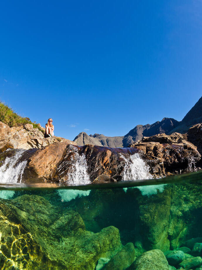 Clear stream with visible underwater rocks, small waterfall, rocky landscape and mountains under a blue sky. Person sits on rocks above the water.