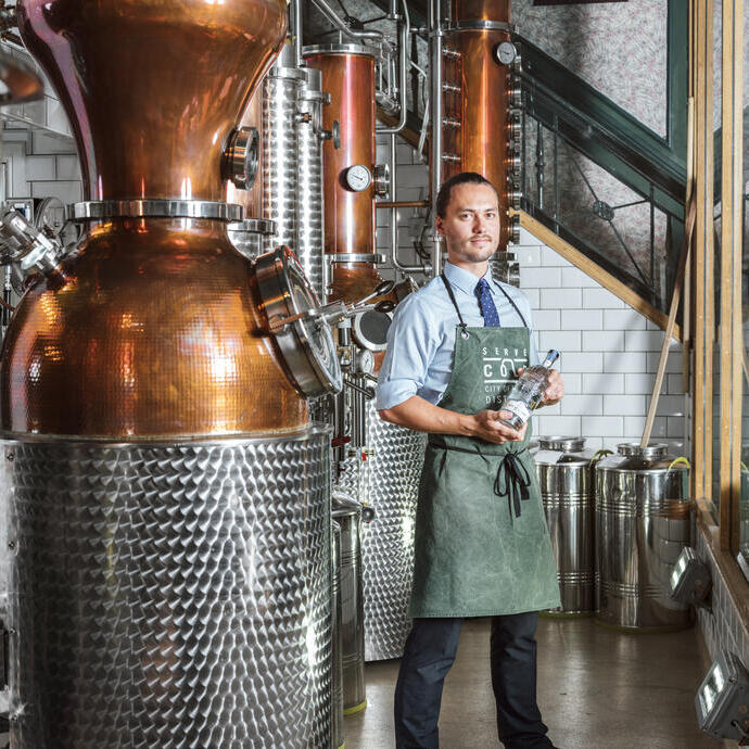Homme portant un tablier vert debout à côté d'un alambic en cuivre dans une distillerie de gin