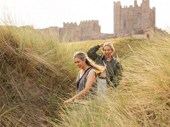 Two women walking in the sand dunes by the sea with a castle in the background