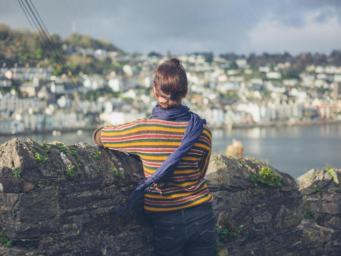 A young woman is standing by a stone wall in a village and is looking at the boats moored in the estuary