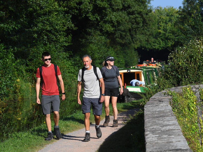 Drei Personen spazieren auf einem Weg entlang eines Kanals mit vertäuten Booten.