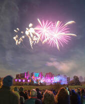 Fireworks display over a ruined castle watched by a crowd of people in the evening