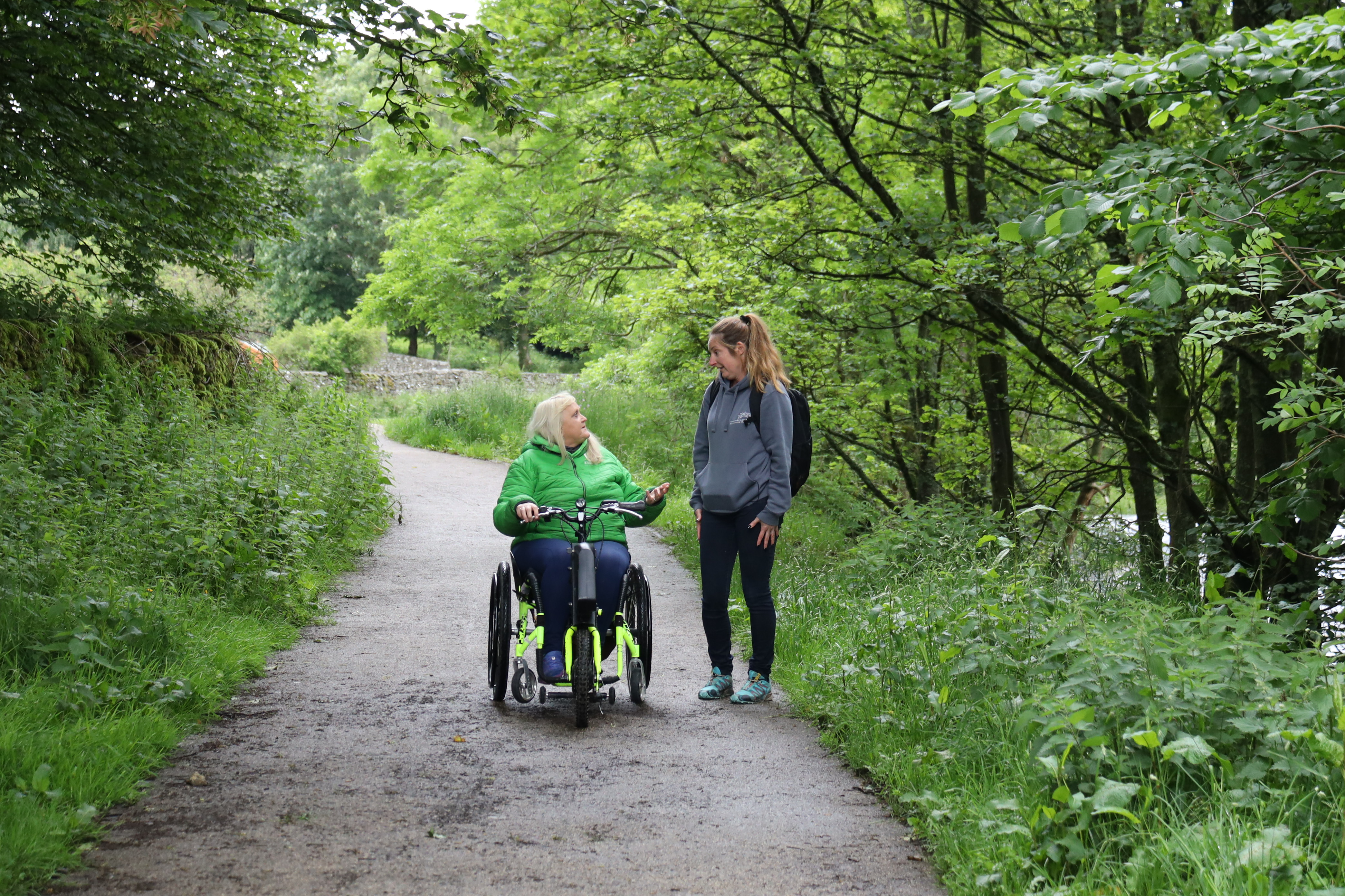 Two women, one on a wheelchair cycle, on a path surrounded by trees