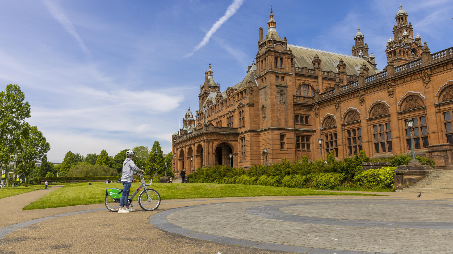 Persona con bicicleta junto a un edificio histórico de ladrillo rojo bajo un cielo azul en un día soleado.