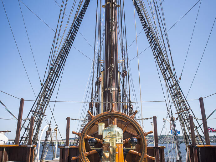 Wooden ship's wheel and rigging on the deck of a historic sailing vessel, with blue sky and seaside background.
