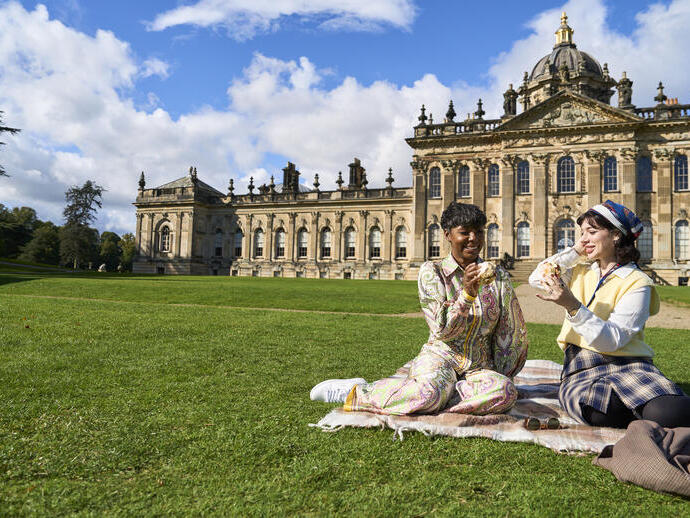 Zwei Personen sitzen auf einer Decke auf dem Rasen vor einem großen historischen Gebäude.