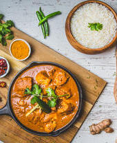 A skillet of chicken curry with rice, surrounded by fresh vegetables and spices on a rustic table.