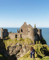 People looking at an historic stony castle from a rocky outcrop cliff over the ocean.