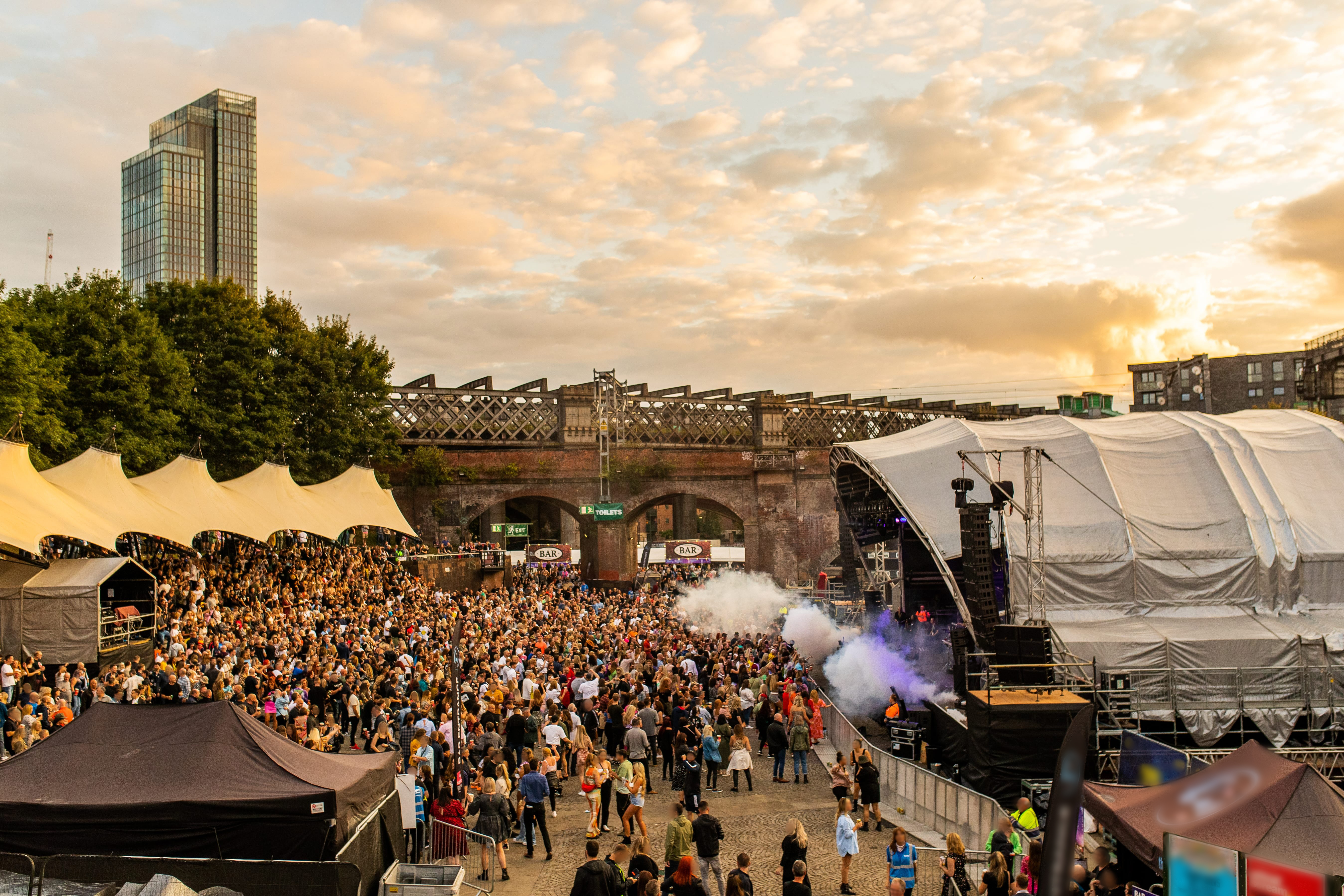 Crowds of people at a festival with stage on right in a city in evening light