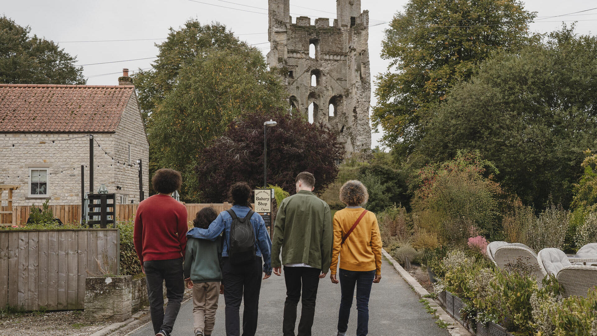 Five people walk towards a stone tower ruin, with a house and garden beside the road. Trees and greenery surround the area.