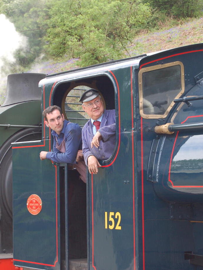 Two men wearing overalls standing in the cab of a vintage steam train.