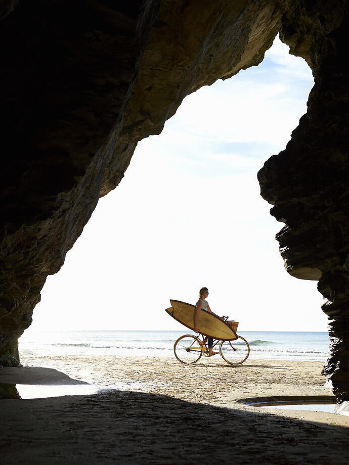 Persona en bicicleta con tabla de surf en la playa, vista desde el interior de una cueva.