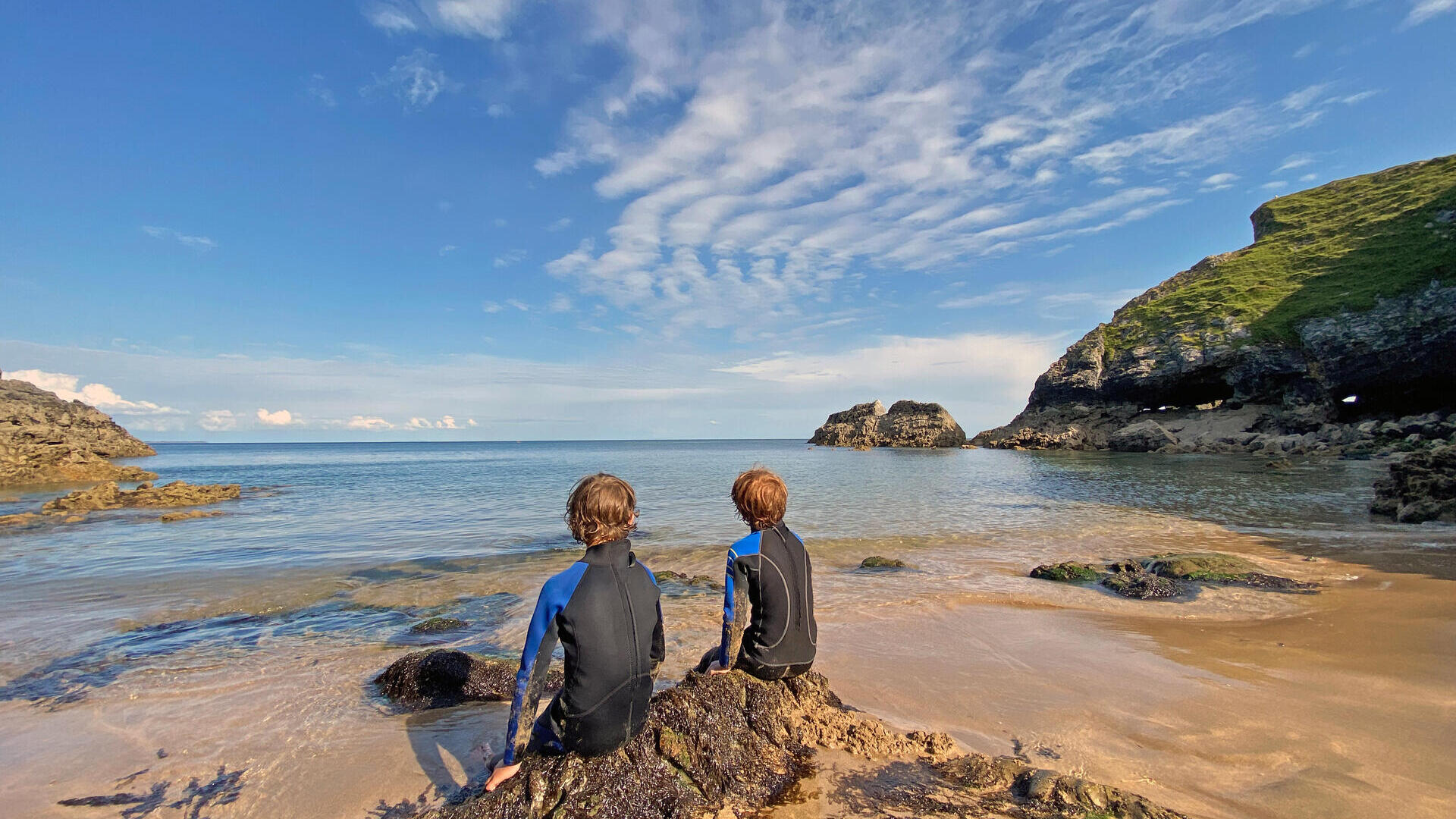 Back view of two young boys in wetsuits sitting on a rock looking out to sea with blue sky