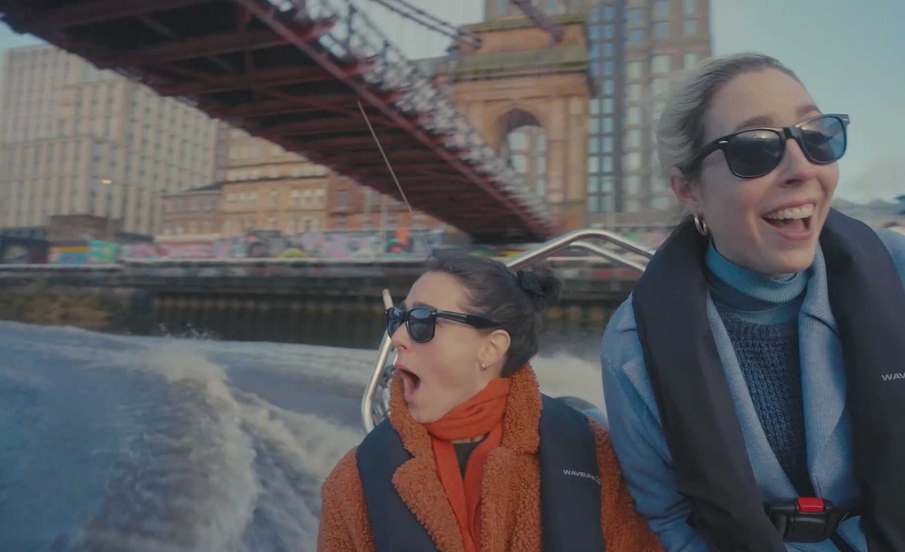 Two women on a motorboat on a river, with a bridge and buildings in the background