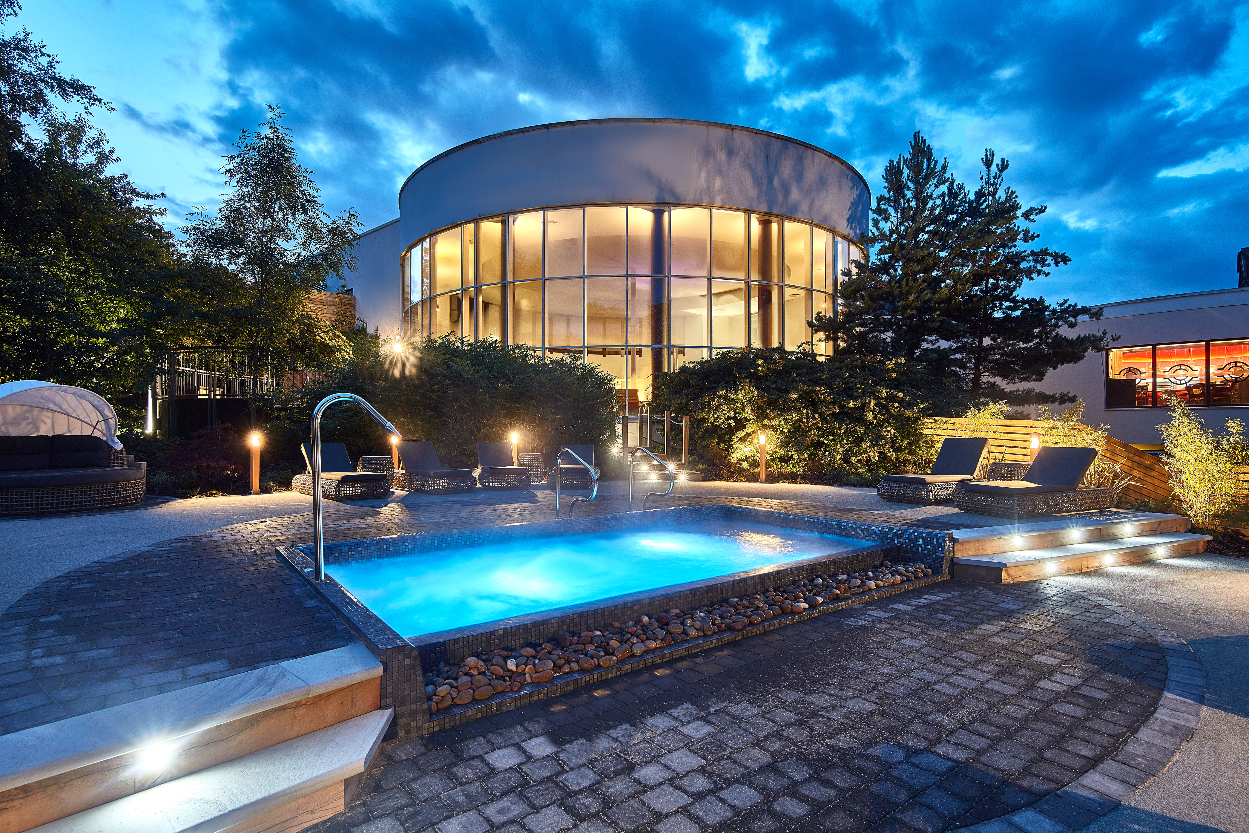 The outdoor spa area at dusk with a lit outdoor spa pool surrounded by loungers and the glass fronted circular spa building in the background. 