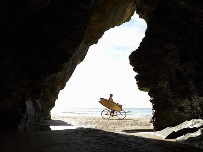 Persona en bicicleta con tabla de surf en la playa, vista desde el interior de una cueva.