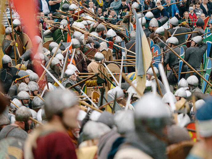 Large crowd of people in historical costumes reenact a medieval battle with shields and spears, watched by an audience.