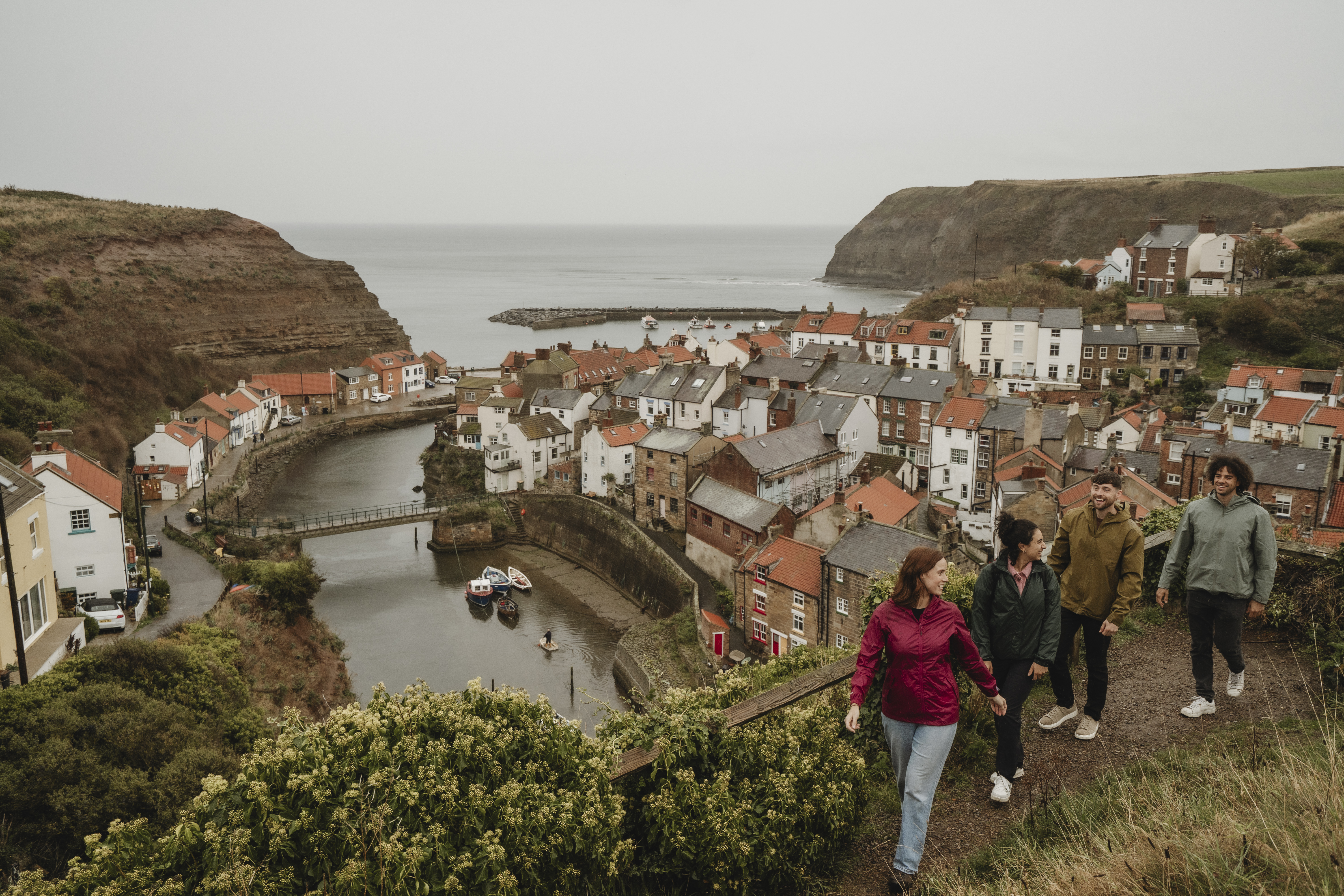 Four people walk along a hillside path overlooking a coastal village with red-roofed houses, a river, boats, and cliffs by the sea.