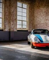 Auto sportiva d'epoca bianca con strisce blu e paraurti rosso in un magazzino di mattoni con grandi finestre.