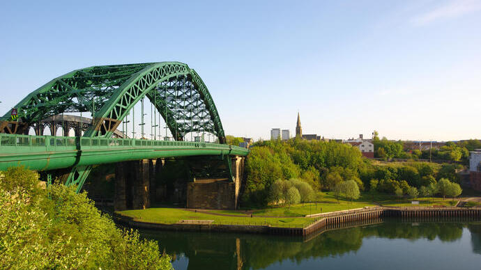 Blick auf eine grüne Eisenbrücke über einen Fluss, umgeben von Bäumen und Gebäuden