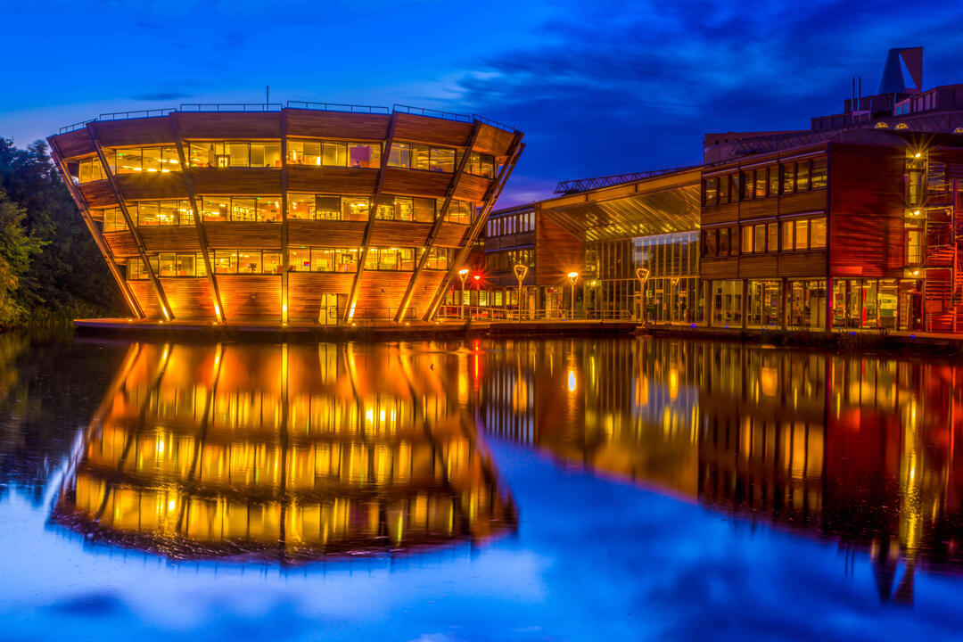 A modern, illuminated building with wooden panels is reflected in a calm body of water at night, against a deep blue sky.