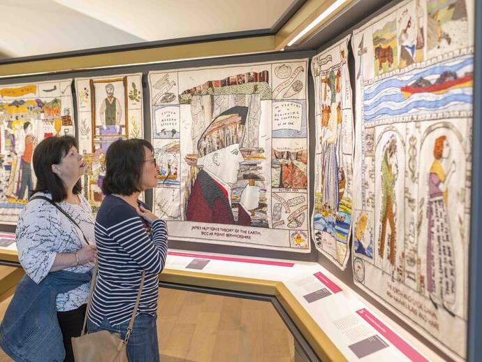 Two people viewing a display of embroidered panels in a museum setting, with illustrated historical and geological themes.
