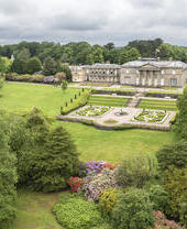 A stately neoclassical mansion with formal gardens, surrounded by lush parkland and dense trees under a cloudy sky.