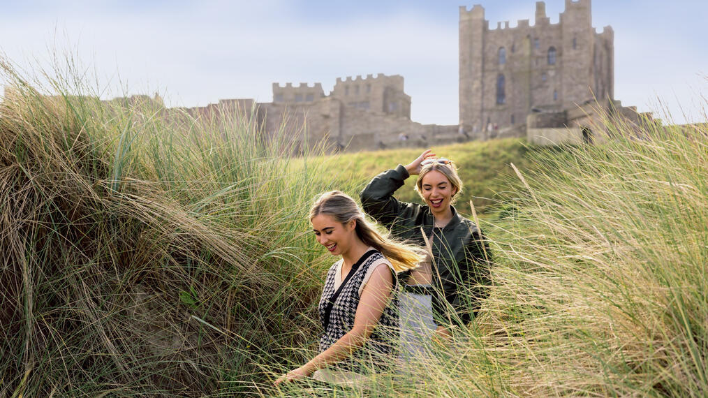 Two women walking in the sand dunes by the sea with a castle in the background