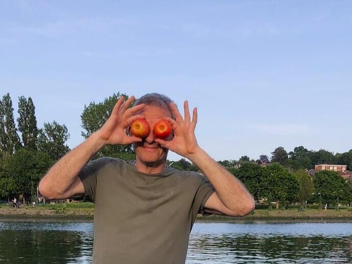 A person stands by the water holding two apples in front of their eyes, with trees and houses in the background.