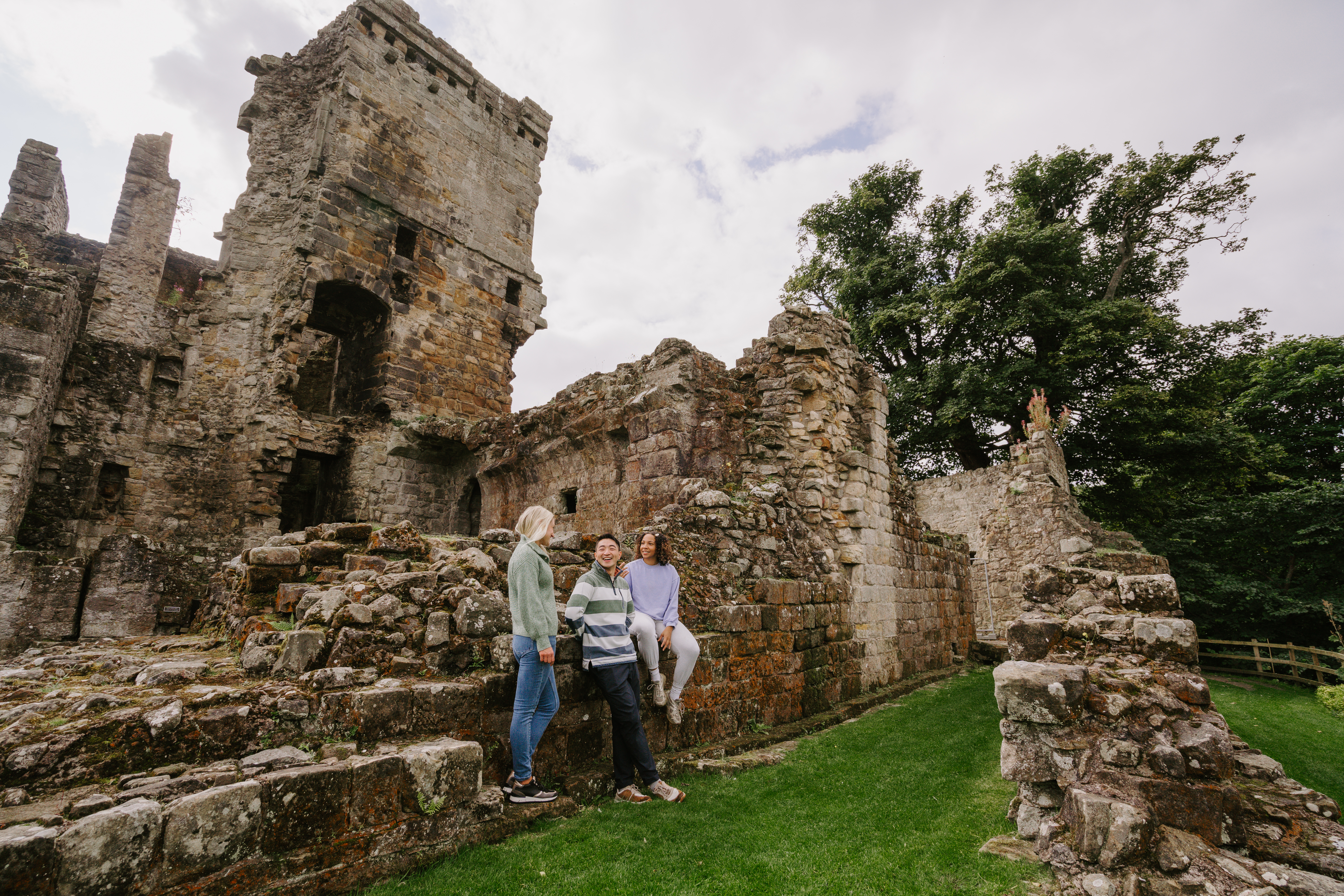Two women and a man sitting on the ruins of a large castle and garden area.