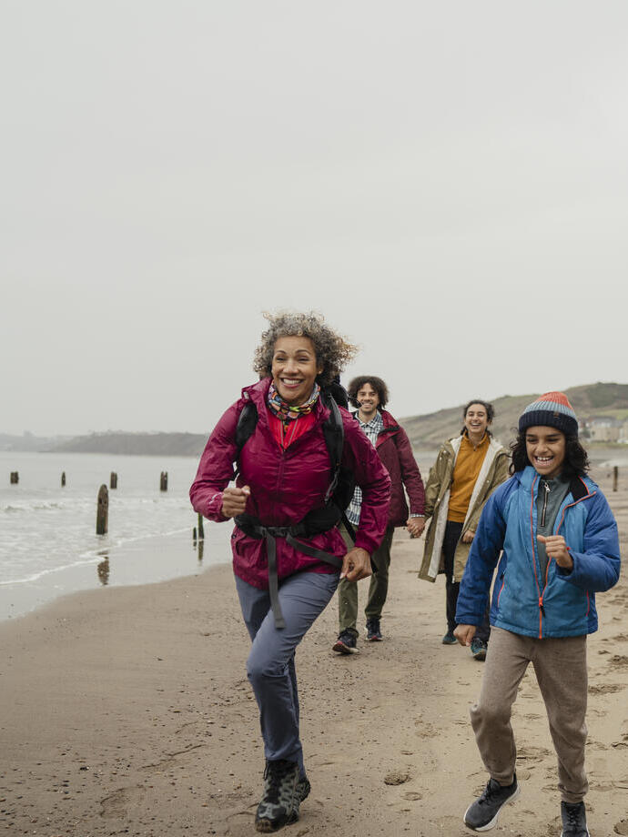 Group of people hiking and smiling along a sandy beach with wooden groynes, some houses, cliffs, and sea in the background on a cloudy day.