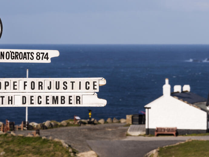 White signpost on a clifftop with the sea and a white building in the distance
