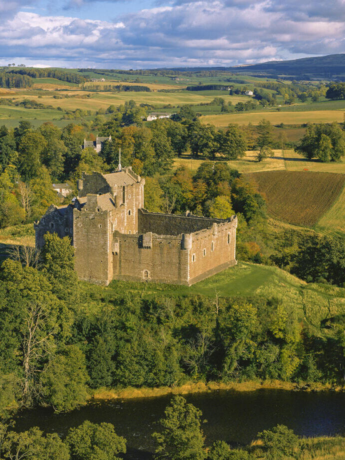 Exterior of a partly ruined medieval castle on a small hilltop overlooking fields and trees in autumn.