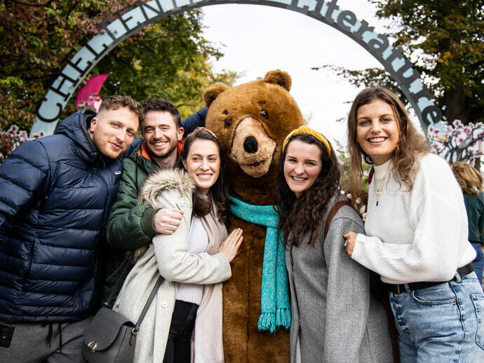 أشخاص يقفون مع دمية دب كبيرة تحت لافتة مقوسة مكتوب عليها "شلتنهام الأدب"