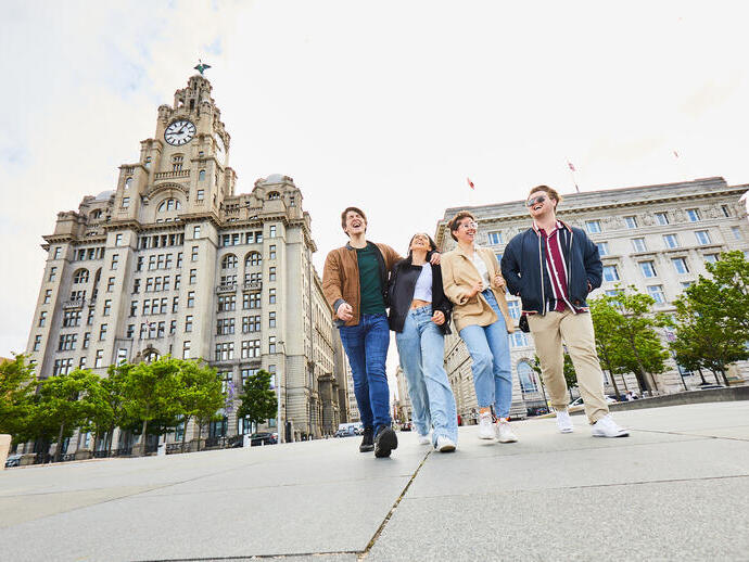 أربعة أصدقاء يستكشفون مدينة محلية وهم يسيرون على طول كورنيش الواجهة البحرية، مع مبانٍ تاريخية في الخلفية