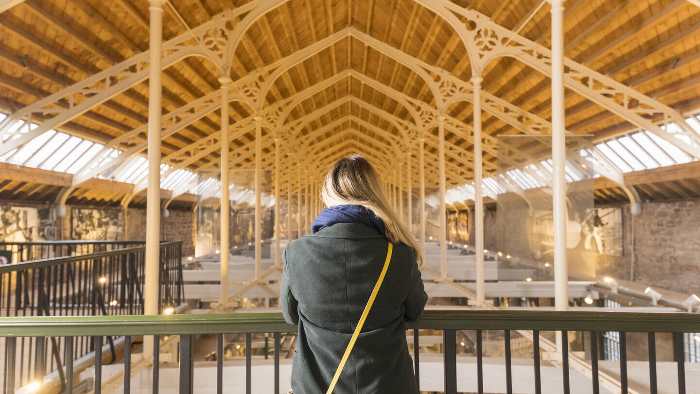Person with a yellow bag stands on a balcony, overlooking a spacious hall with a high wooden ceiling and glass windows.