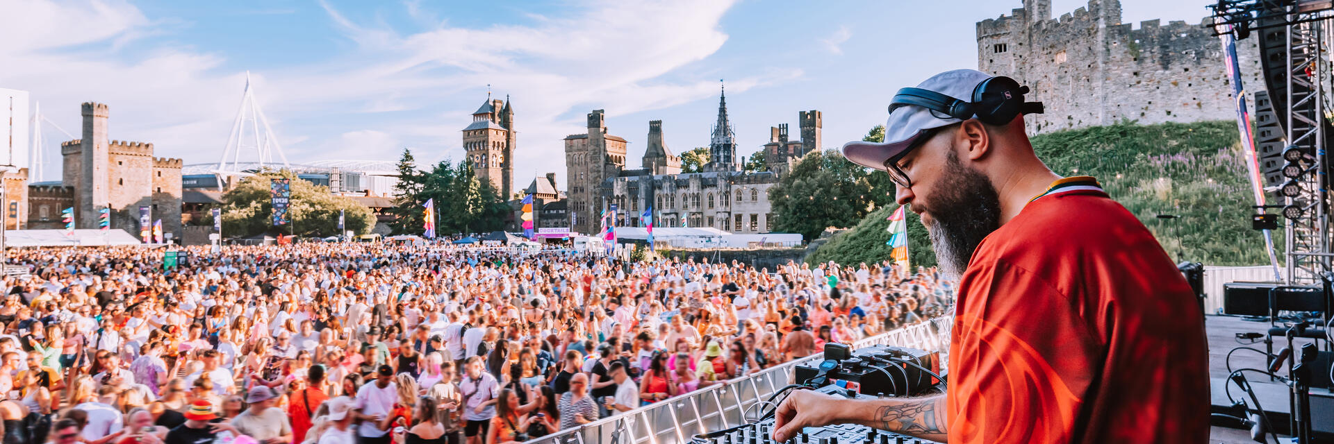 DJ spielt vor großem Publikum auf einem Musikfestival im Freien, mit einer Burg im Hintergrund und blauem Himmel.