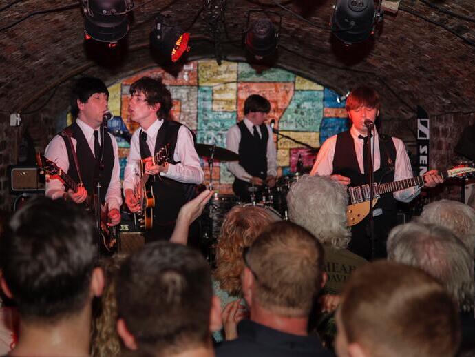 A band dressed in matching black vests and ties performs live in a crowded, arched brick venue with a colorful graffiti-style wall in the background.