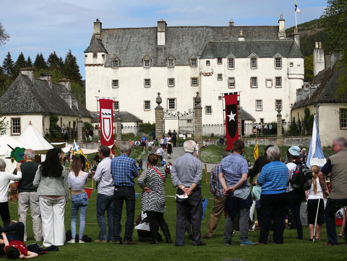A crowd enjoys an outdoor event in front of a large historic castle, with medieval banners and tents on the grounds.
