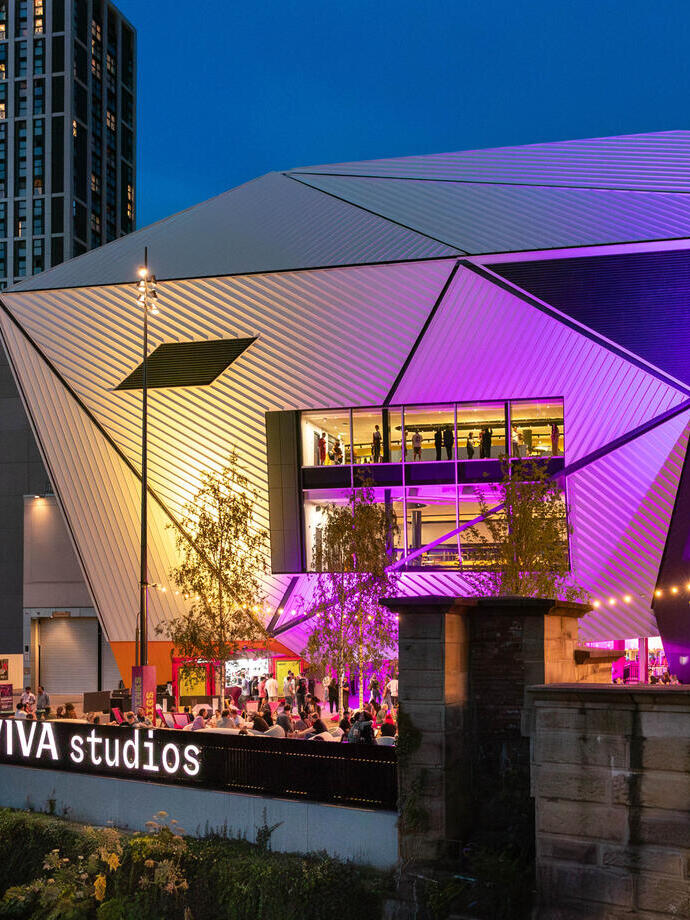 Bâtiment moderne et géométrique avec des panneaux métalliques éclairés en violet la nuit, intitulé AVIVA studios, entouré d'arbres et d'immeubles.